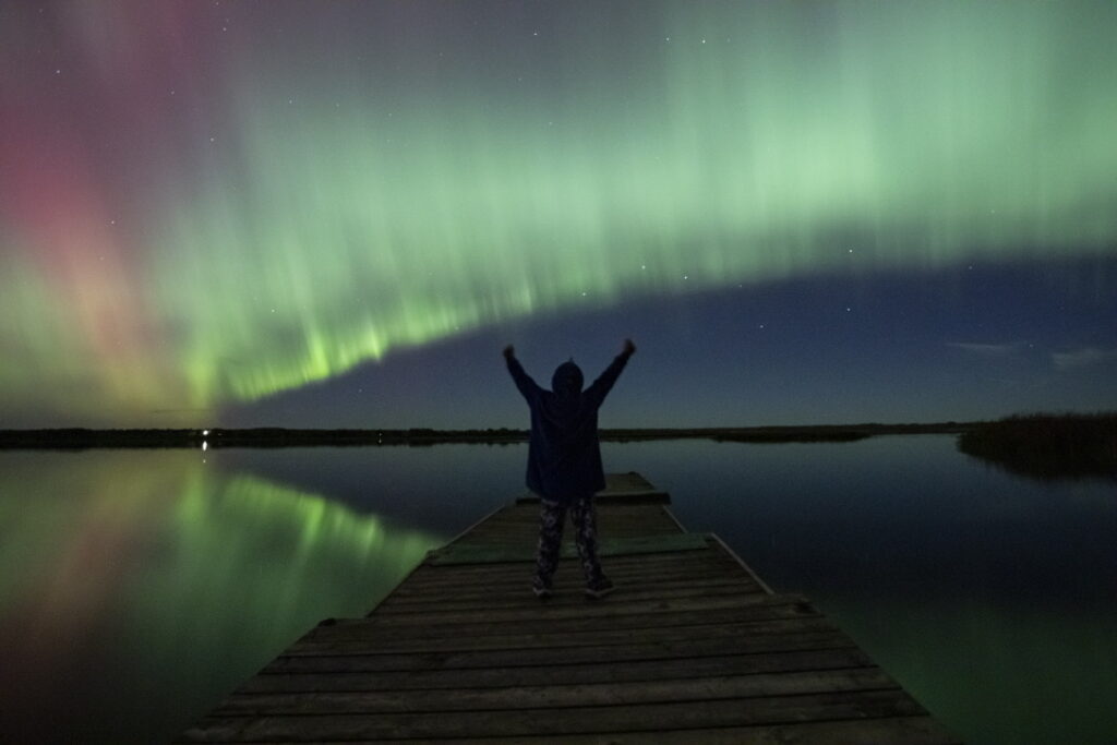 Northern lights reflect off the water while a boy stands on a wharf.