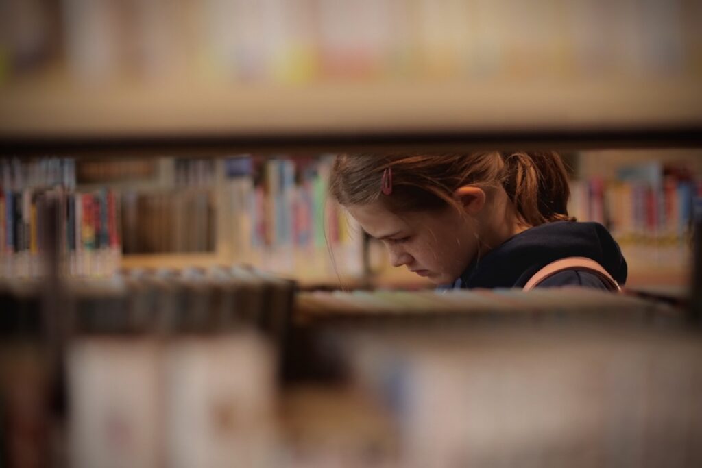Young girl is seen through the shelves of books at a library.