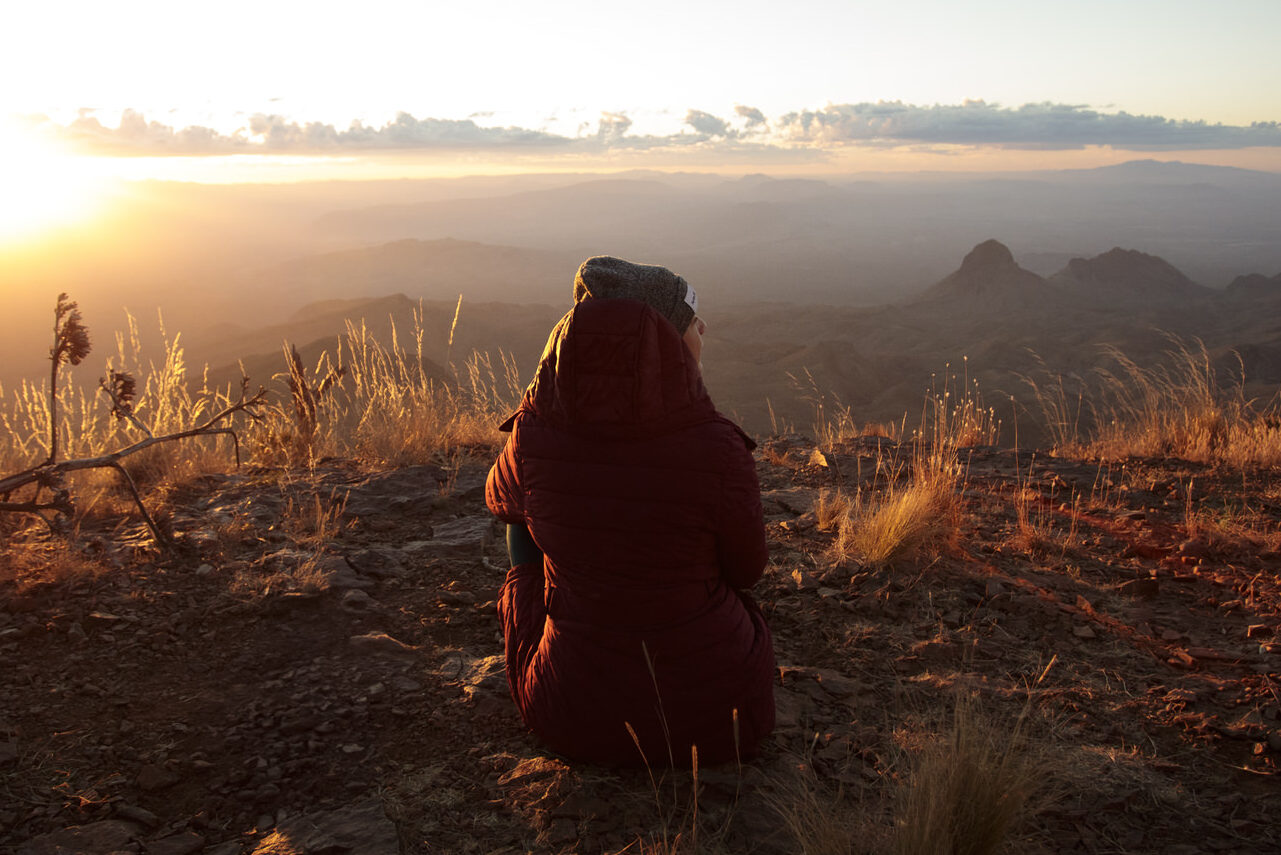 Ali sits on a cliffside overlooking mountains below at sunrise. Big Bend National Park.