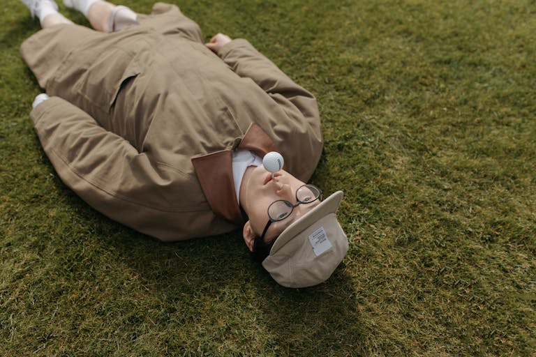 Fashionable man with golf ball on cap, lying on grass, showcasing a creative and quirky golf concept.
