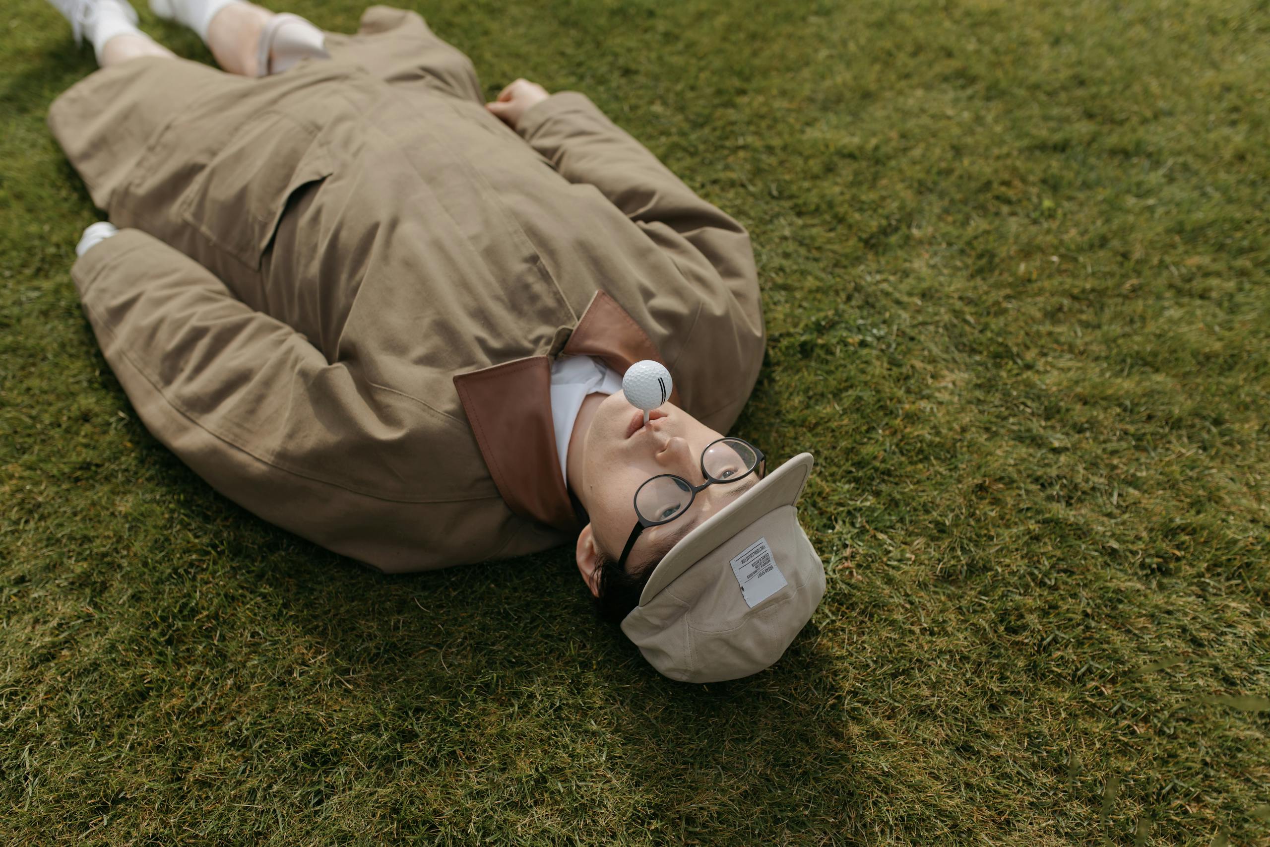 Fashionable man with golf ball on cap, lying on grass, showcasing a creative and quirky golf concept.